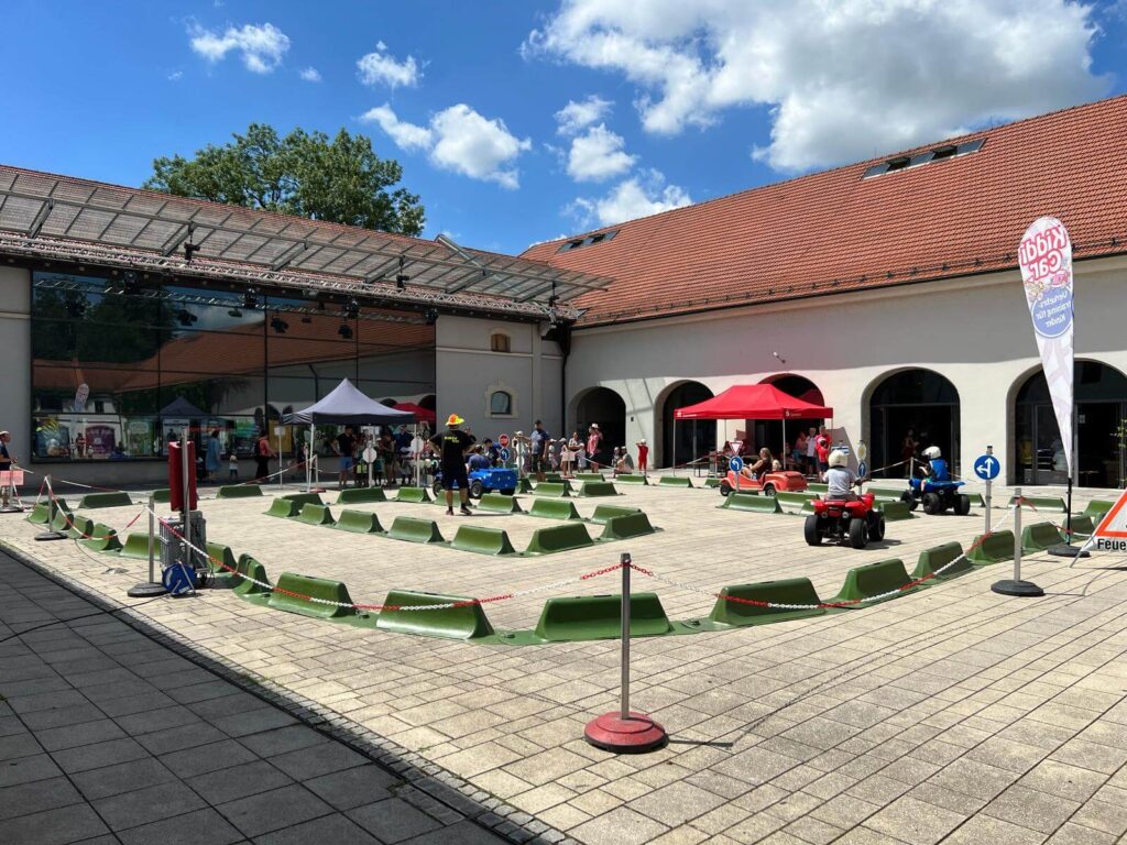 Kiddi-Car Mitarbeiter mit gelbem Sonnenhut beaufsichtigt die Kinder beim sicheren Fahren auf dem KNAX-Fest 2025 im Kloster Fürstenfeld in Fürstenfeldbruck.

